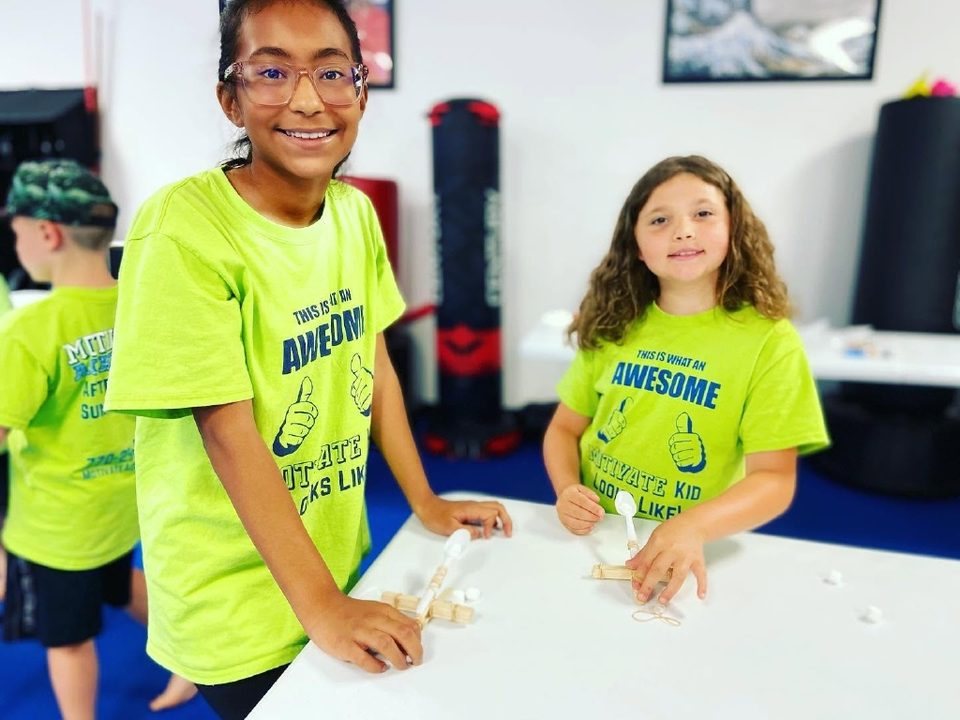 Two smiling girls in Motivate shirts building popsicle-stick catapults at a craft table