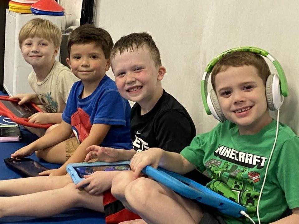 Four smiling boys sitting against a wall playing on tablets, one wearing green headphones