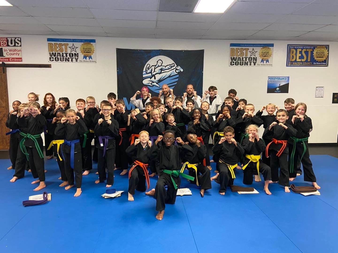 A big group of Motivate kids in black uniforms and colored belts posing with their fists up on the blue mats
