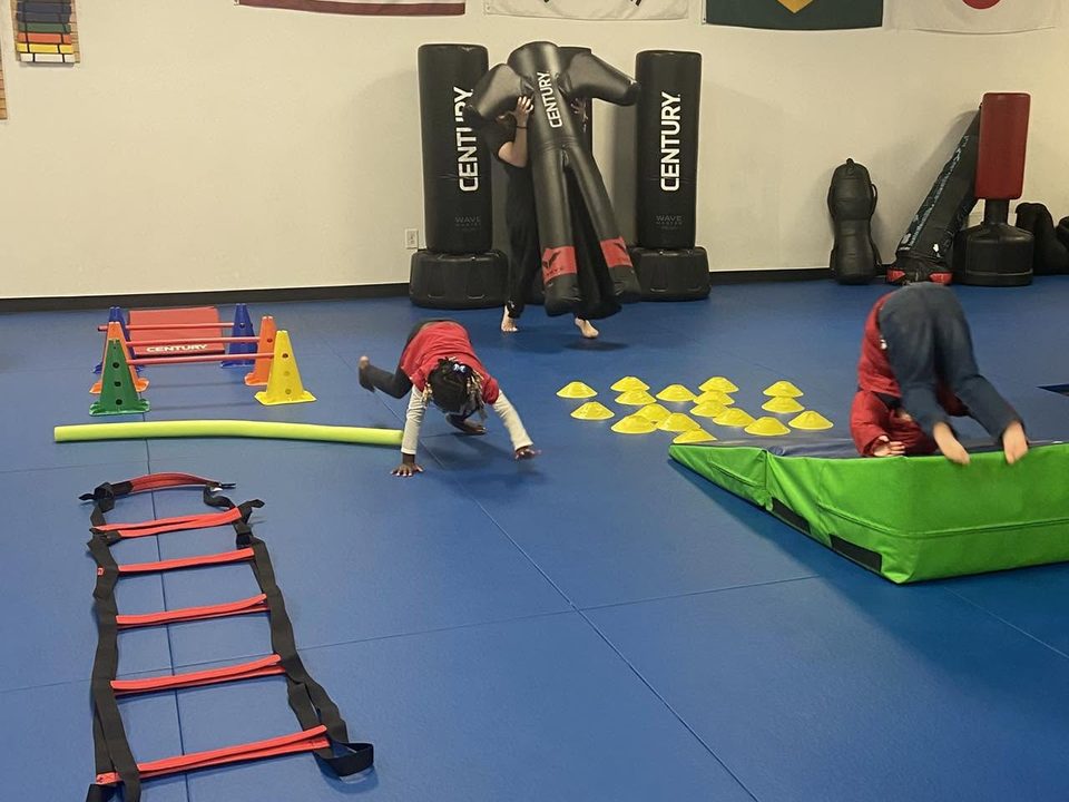 Kids rolling and crawling through an obstacle course with cones, hurdles, an agility ladder and a green tumbling mat